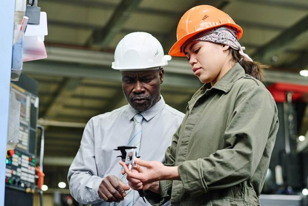 Two manufacturing workers inspect a metal component together on a factory floor.
